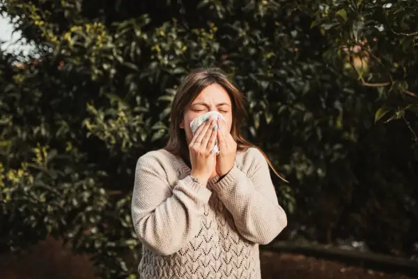 Kobieta w beżowym swetrze chusteczką wyciera nos na tle zielonych liści. Wiosna, na co teraz alergia?