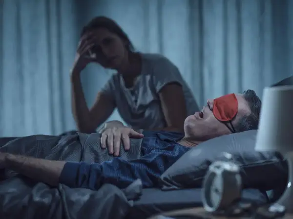 Zestresowana kobieta szuka sposobów na chrapanie swojego partnera, który głośno śpi w czerwonej opasce na oczy.